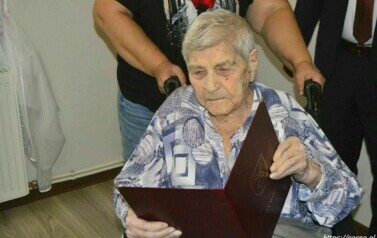 Jubilatka siedząca na w&oacute;zku dla os&oacute;b z niepełnosprawnościami czyta list gratulacyjny od W&oacute;jta Gminy Sośno.