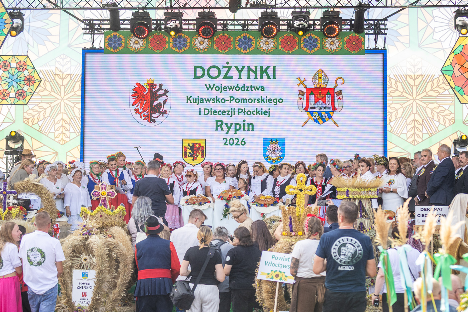 Zdjęcie przedstawia uroczyste obchody Dożynek Wojew&oacute;dztwa Kujawsko-Pomorskiego i&nbsp;Diecezji Płockiej w&nbsp;Rypinie w&nbsp;2026 roku. Na scenie stoi liczna grupa uczestnik&oacute;w w&nbsp;tradycyjnych strojach ludowych i&nbsp;odświętnych ubraniach, prezentujących okazałe wieńce dożynkowe oraz&nbsp;bochny chleba. W tle widoczny jest ekran z&nbsp;napisem &bdquo;Dożynki Wojew&oacute;dztwa Kujawsko-Pomorskiego i&nbsp;Diecezji Płockiej, Rypin 2026&rdquo;, a&nbsp;przed sceną zgromadzili się mieszkańcy i&nbsp;uczestnicy uroczystości.
