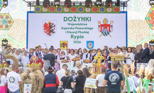 Zdjęcie przedstawia uroczyste obchody Dożynek Województwa Kujawsko-Pomorskiego i Diecezji Płockiej w Rypinie w 2026 roku. Na scenie stoi liczna grupa uczestników w tradycyjnych strojach ludowych i odświętnych ubraniach, prezentujących okazałe wieńce dożyn