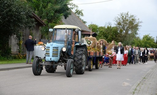 Korowód dożynkowy ulicą Tonina – na pierwszym planie zabytkowy niebieski traktor z przyczepą ozdobioną wieńcami i dekoracjami z plonów, za nim mieszkańcy i delegacje dożynkowe. W tle widoczne dorożki oraz zabudowania i zieleń.
