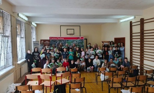 Grupowe zdjęcie uczniów Szkoły Podstawowej im. Janusza Korczaka w Sośnie oraz zaproszonych gości podczas wręczania kasków rowerowych. Uczniowie trzymają otrzymane kaski, a w tle widoczna jest plansza z nazwą Gminy Sośno.