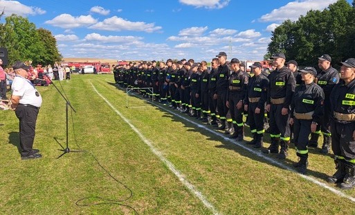 Na boisku w Sośnie podczas Gminnych Zawodów Pożarniczych stoi w szeregu kilkudziesięciu strażaków z jednostek OSP, ubranych w czarne stroje strażackie. Przed nimi, przy mikrofonie, przemawia prowadzący zawody. W tle widoczne są wozy strażackie, uczestnicy wydarzenia oraz drzewa i pola. Pogodny dzień z błękitnym niebem i chmurami.
