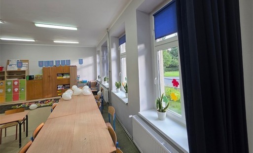 Zdjęcie przedstawia wnętrze sali lekcyjnej w jednej ze szkół podstawowych Gminy Sośno. Widać długi stół, krzesła, szafki z materiałami edukacyjnymi oraz okna z widokiem na zieleń. 