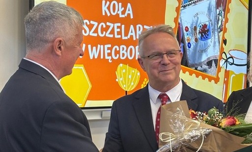 Na zdjęciu widoczny Wójt Gminy Sośno, wręczający bukiet kwiatów. W tle wyświetla się plakat 80lecia Koła Pszczelarzy w Więcborku.